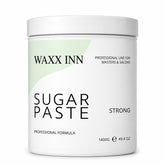 A white plastic jar of WAXX INN Sugar Paste Strong professional formula, featuring a minimalist label with a light green abstract shape and black text, set against a plain white background.